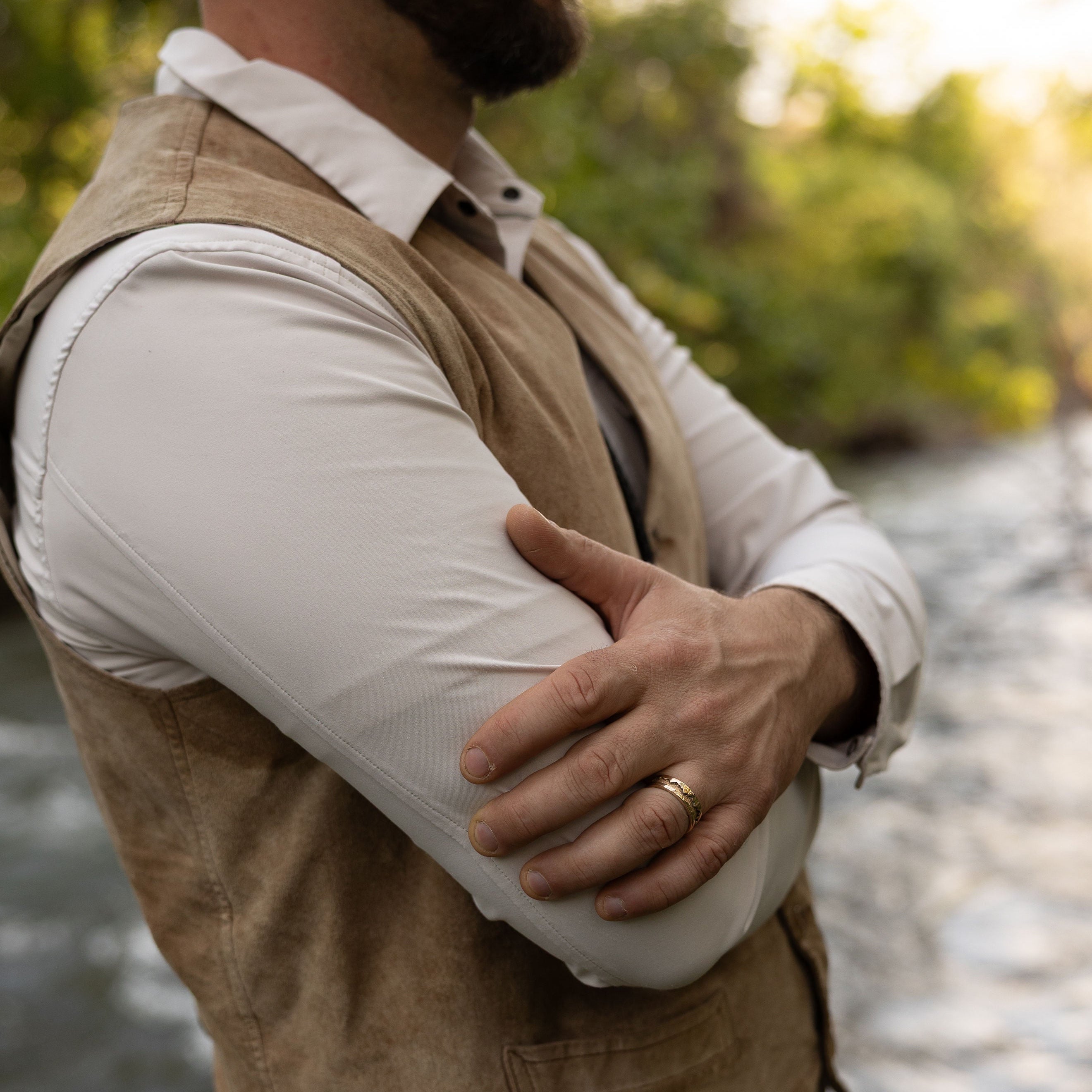 Man wearing a beige vest and white shirt by a river with trees in the background