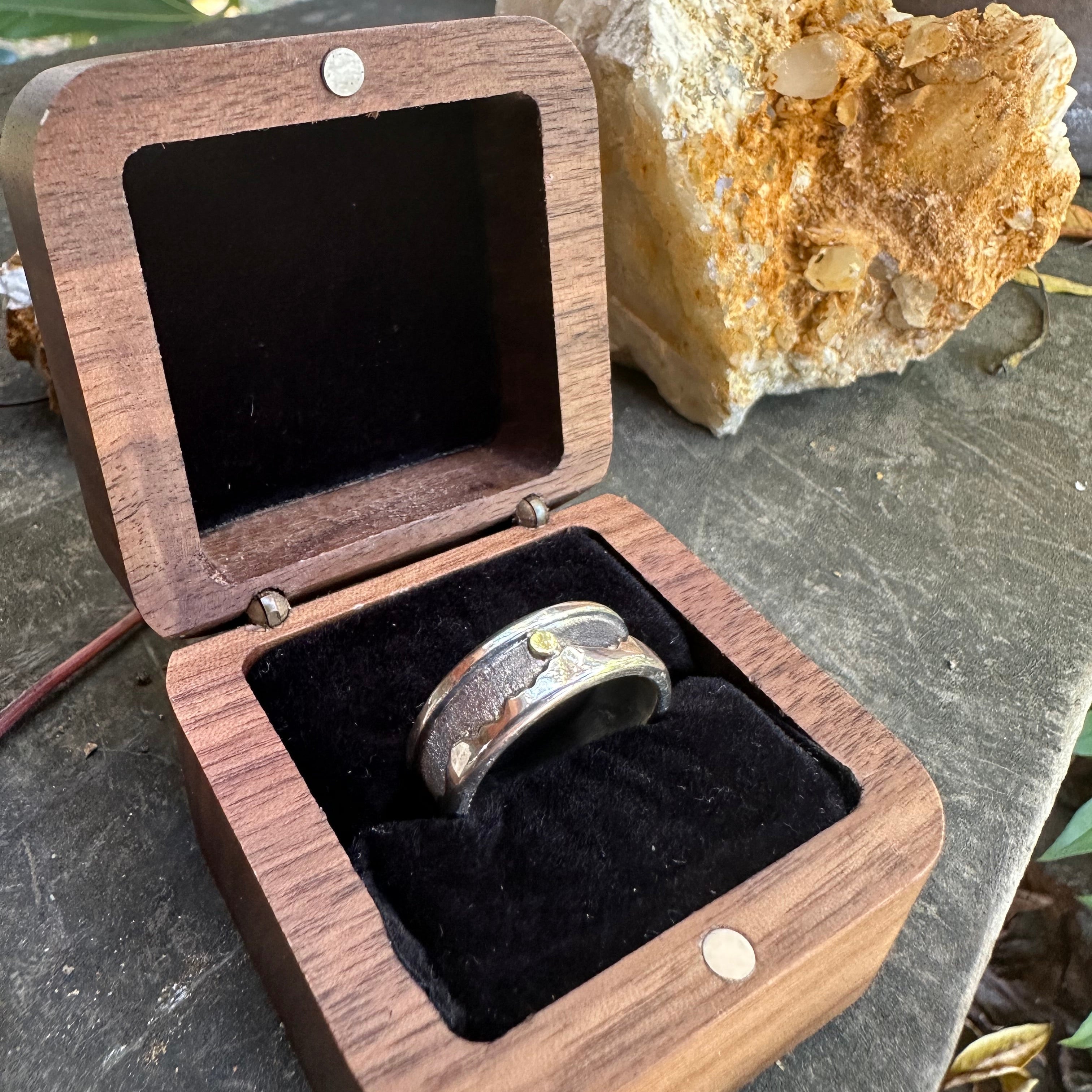 Wooden jewelry box with a silver ring inside, placed on a stone surface with a rock and plant in the background.