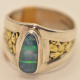 A silver ring featuring a central teardrop-shaped blue-green opal stone with green flashes, flanked by textured yellow-gold inlay details on a light beige background.
