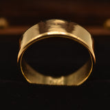 A wide, polished gold band ring displayed on a black velvet jewelry stand against a dark wooden background.