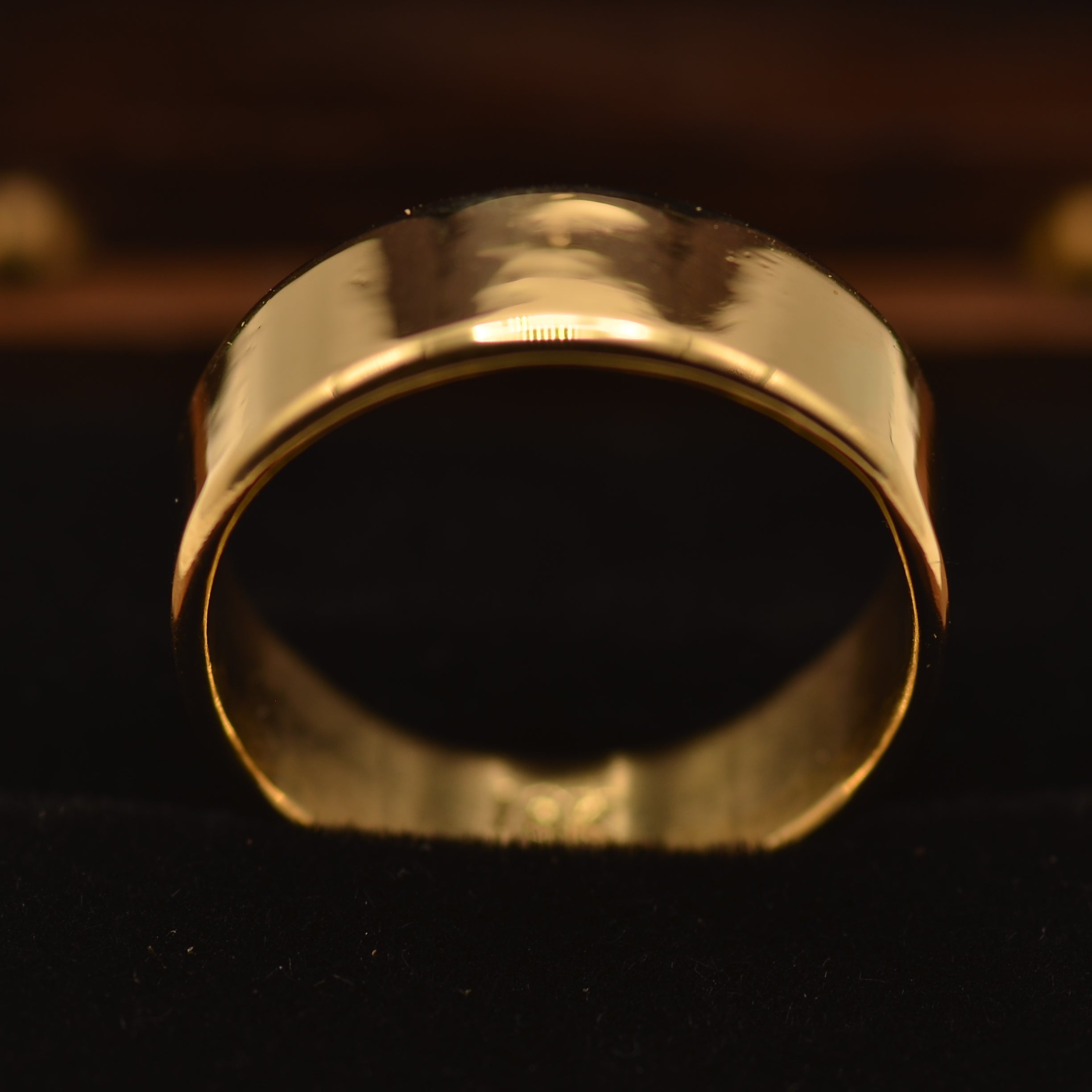 A wide, polished gold band ring displayed on a black velvet jewelry stand against a dark wooden background.