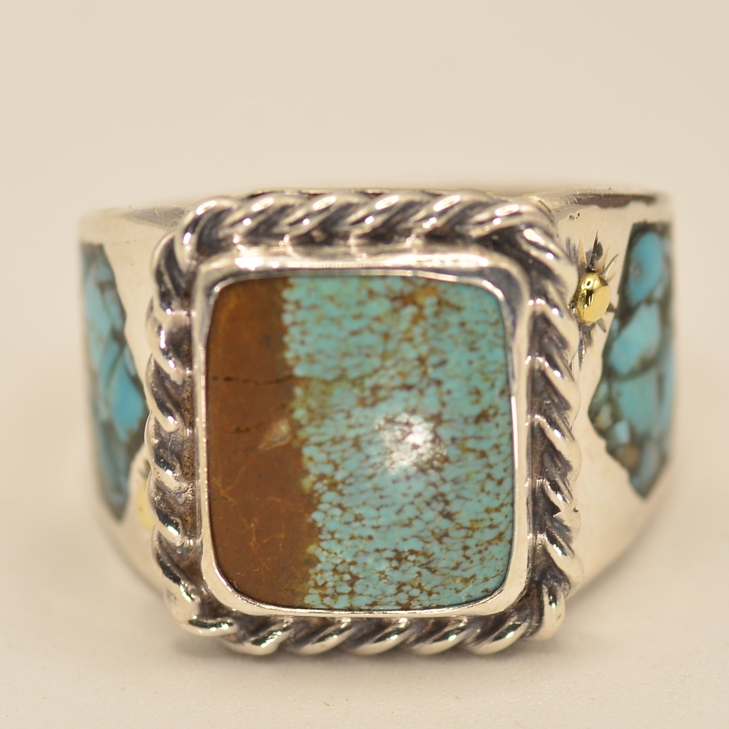 A silver ring featuring a square bezel set with a split center design of brown and turquoise, flanked by small turquoise inlays on a cream background.
