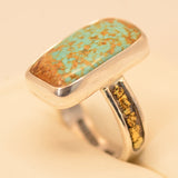 A sterling silver ring featuring a large, rectangular turquoise stone with prominent brown matrix, and a shank decorated with small gold inclusions, displayed on a cream-colored surface.