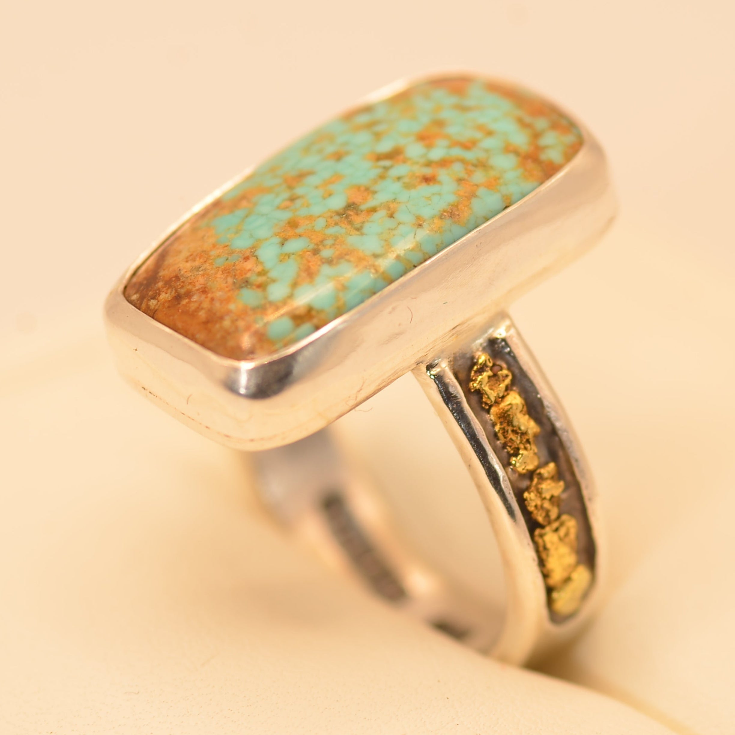 A sterling silver ring featuring a large, rectangular turquoise stone with prominent brown matrix, and a shank decorated with small gold inclusions, displayed on a cream-colored surface.