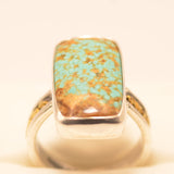 A sterling silver ring featuring a large, rectangular turquoise stone with prominent golden-brown matrix, displayed on a cream-colored ring stand.