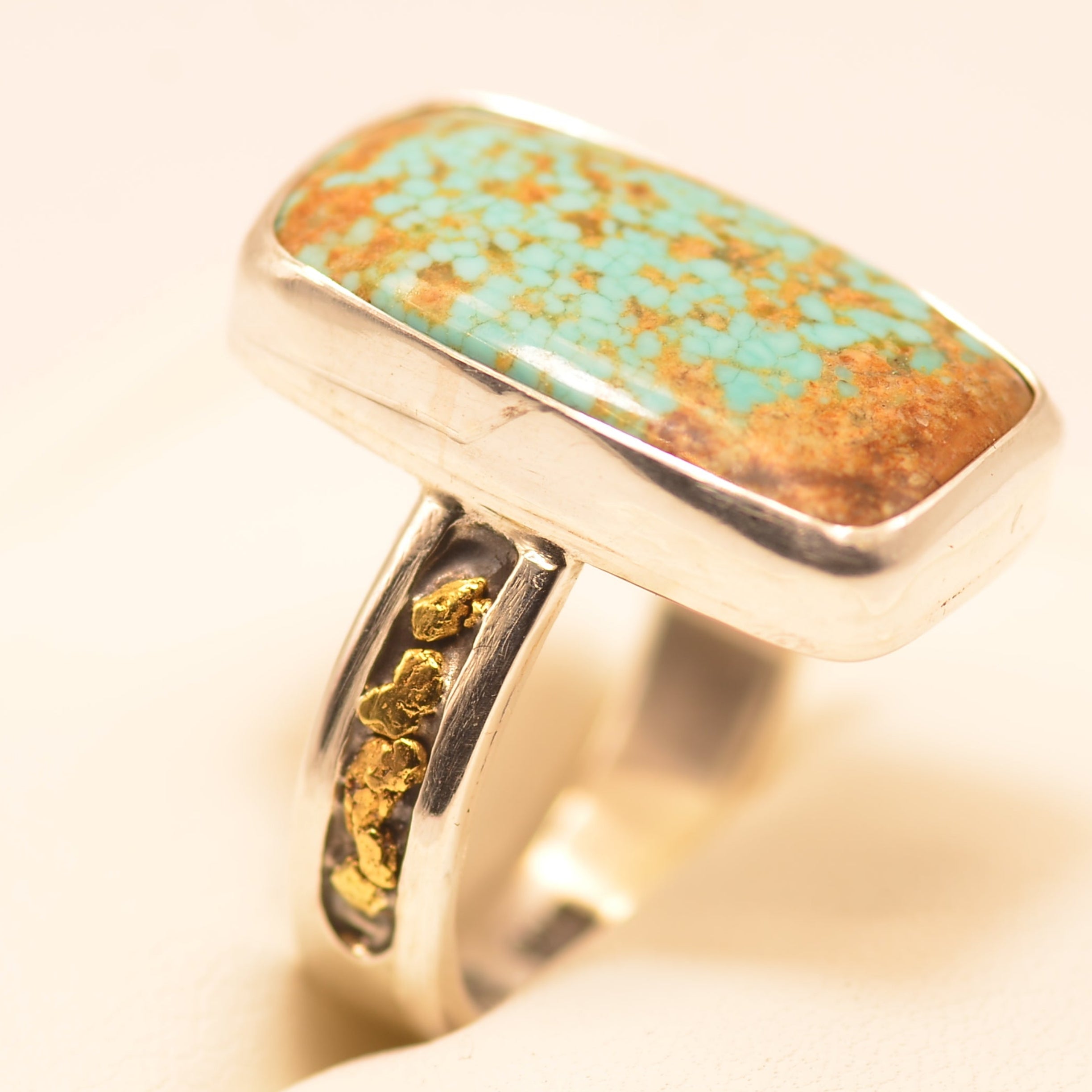 A sterling silver ring featuring a rectangular turquoise stone with prominent coppery brown matrix, displayed on a soft cream-colored surface.
