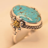 A sterling silver ring featuring an oval turquoise cabochon stone with natural matrix veining, accented by a small yellow gold skull detail on the band, set against a warm beige background.