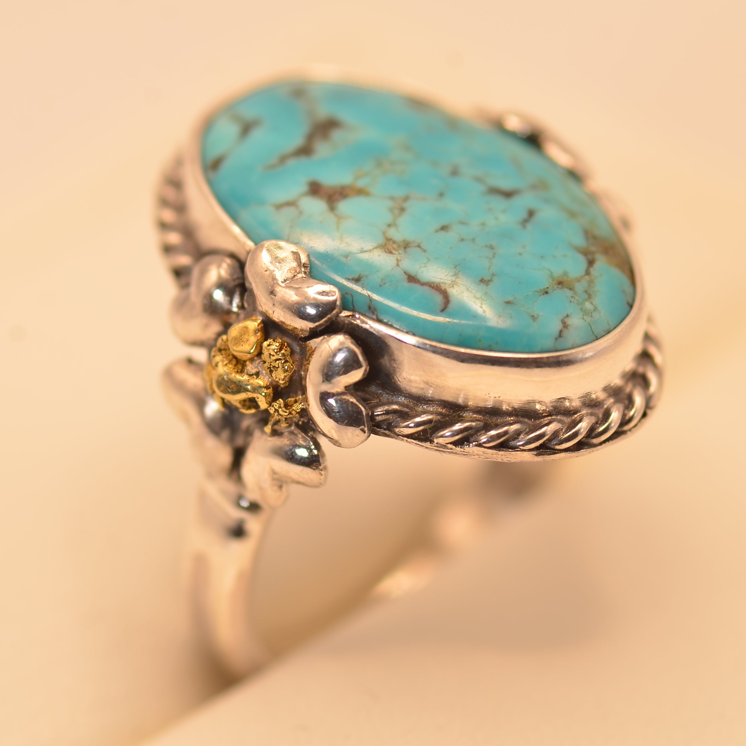 A sterling silver ring featuring an oval turquoise cabochon stone with natural matrix veining, accented by a small yellow gold skull detail on the band, set against a warm beige background.