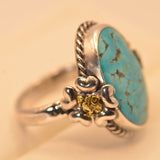 A sterling silver ring featuring a large oval turquoise stone with dark matrix veining, accented by a small yellow gold flower detail on the band, set against a warm beige background.