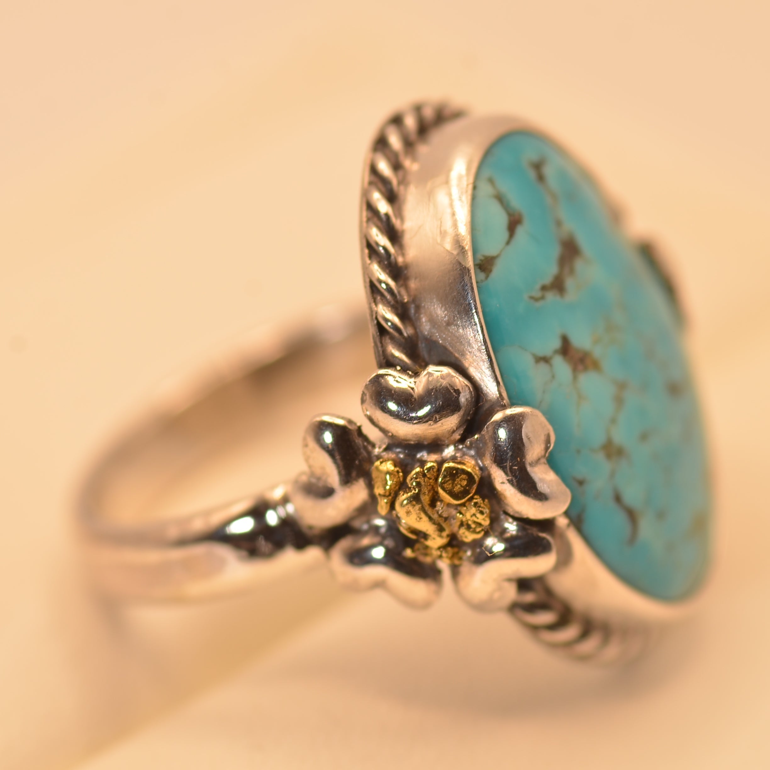 A sterling silver ring featuring a large oval turquoise stone with dark matrix veining, accented by a small yellow gold flower detail on the band, set against a warm beige background.