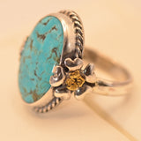 A sterling silver ring featuring a large oval turquoise stone with natural matrix veining, accented by a small gold flower detail on the band, set against a warm cream background.
