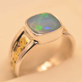 A silver ring featuring a square cabochon opal gemstone with vibrant green and blue flashes, set against a warm beige background.