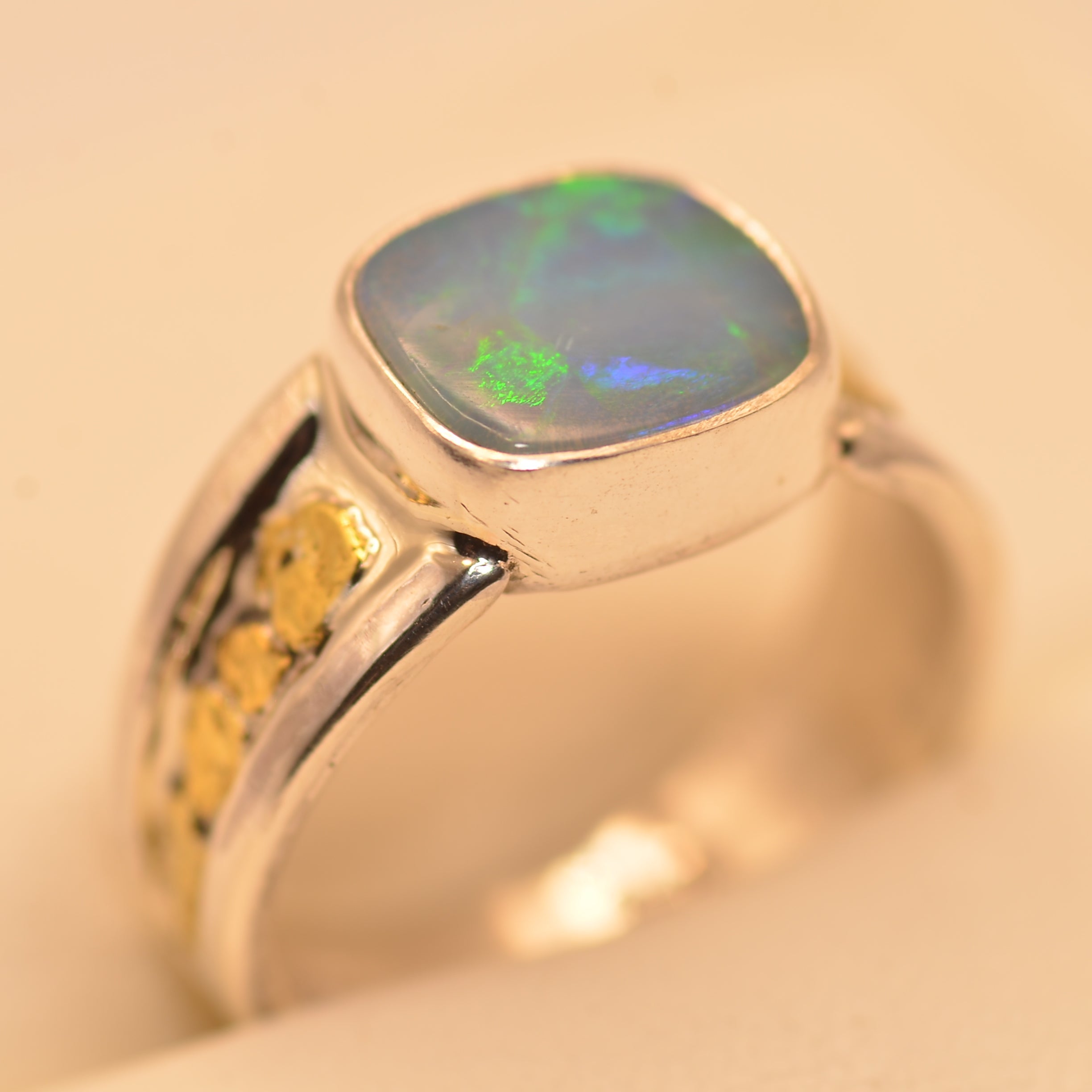 A silver ring featuring a square cabochon opal gemstone with vibrant green and blue flashes, set against a warm beige background.