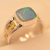 A silver ring featuring a square cabochon opal gemstone with vibrant green and blue play-of-color, set against a warm beige background.