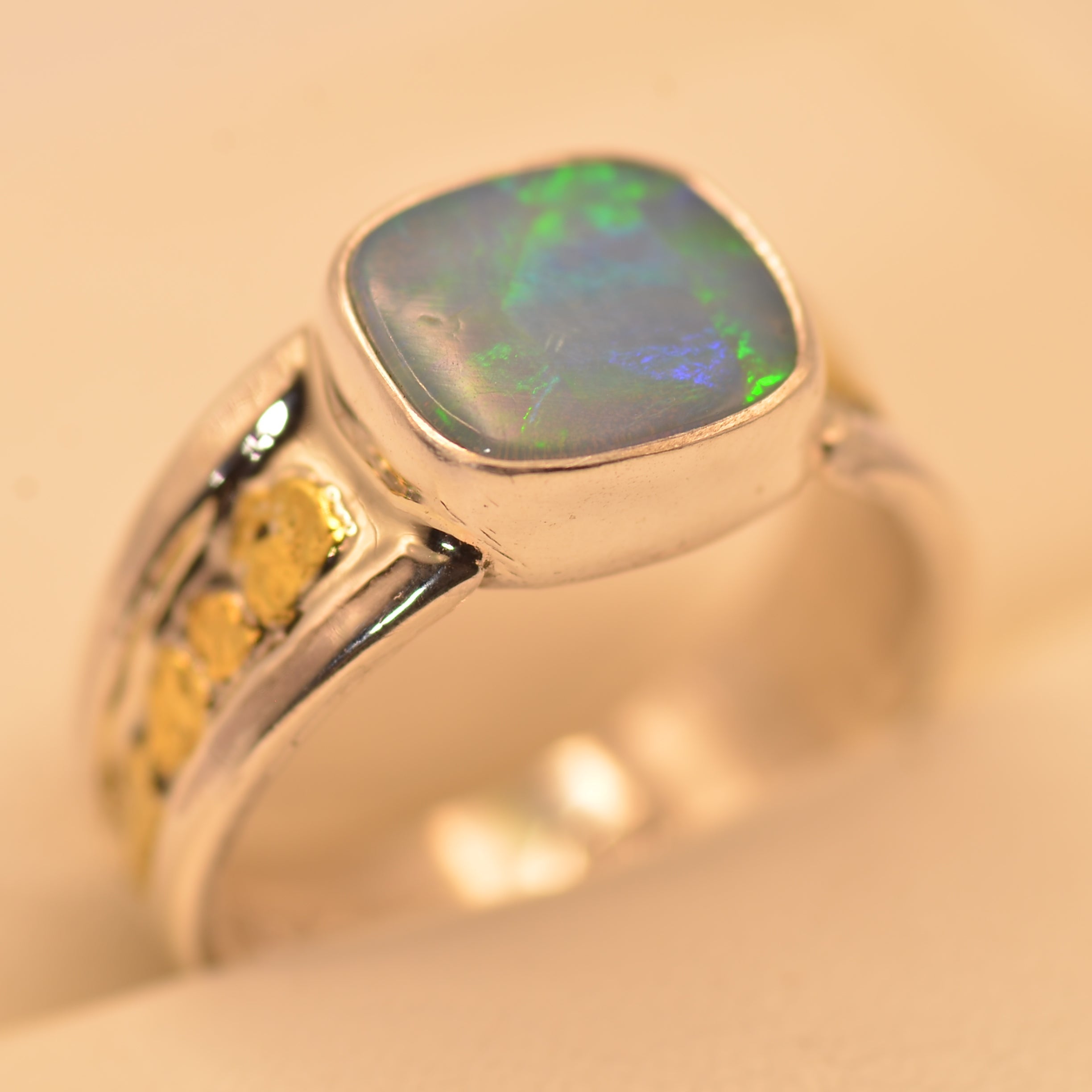 A silver ring featuring a square cabochon opal gemstone with vibrant green and blue play-of-color, set against a warm beige background.