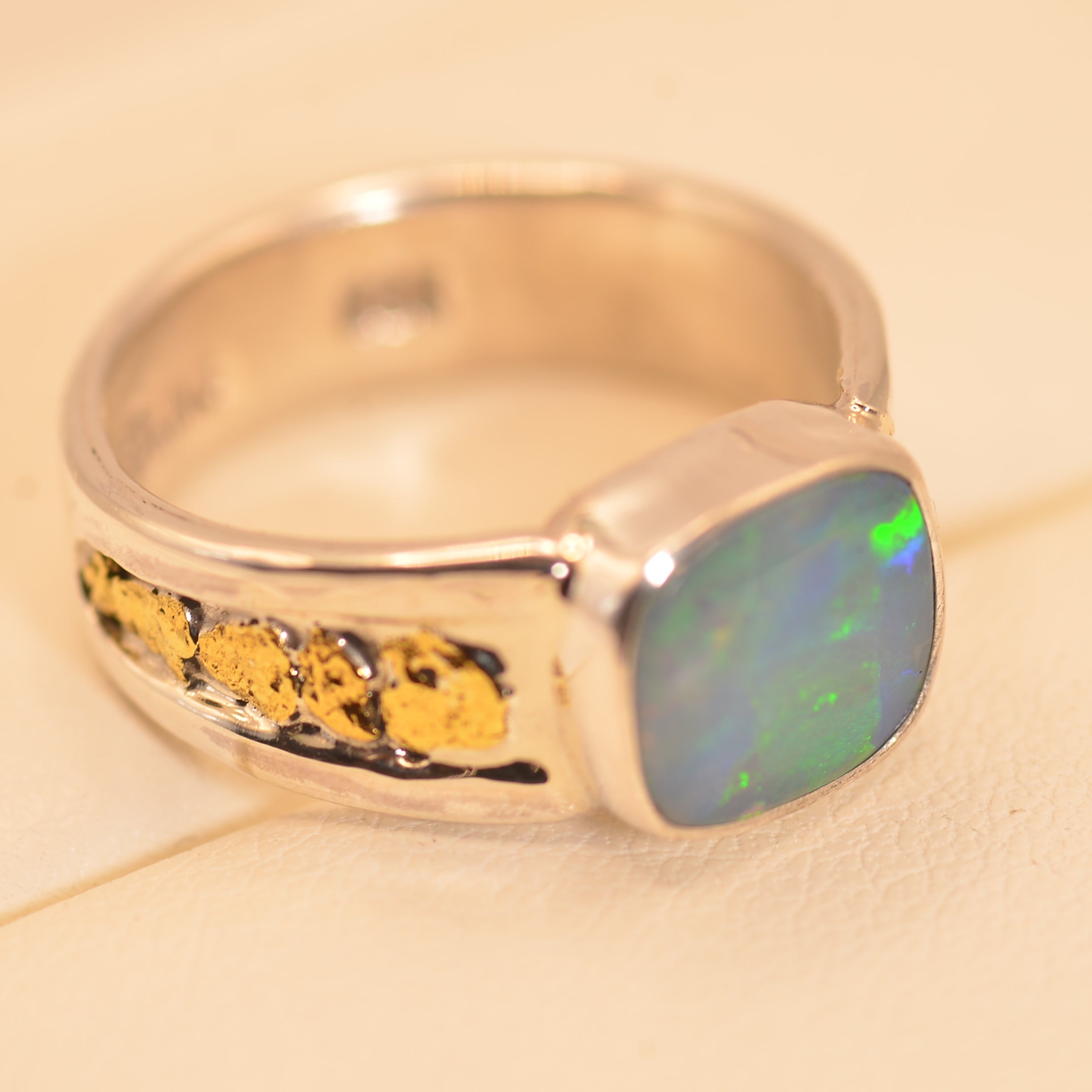A silver band ring featuring a square cabochon opal gemstone with iridescent blue, green, and purple flashes, accented by a row of small yellow gold inlays on the side, set against a warm cream background.