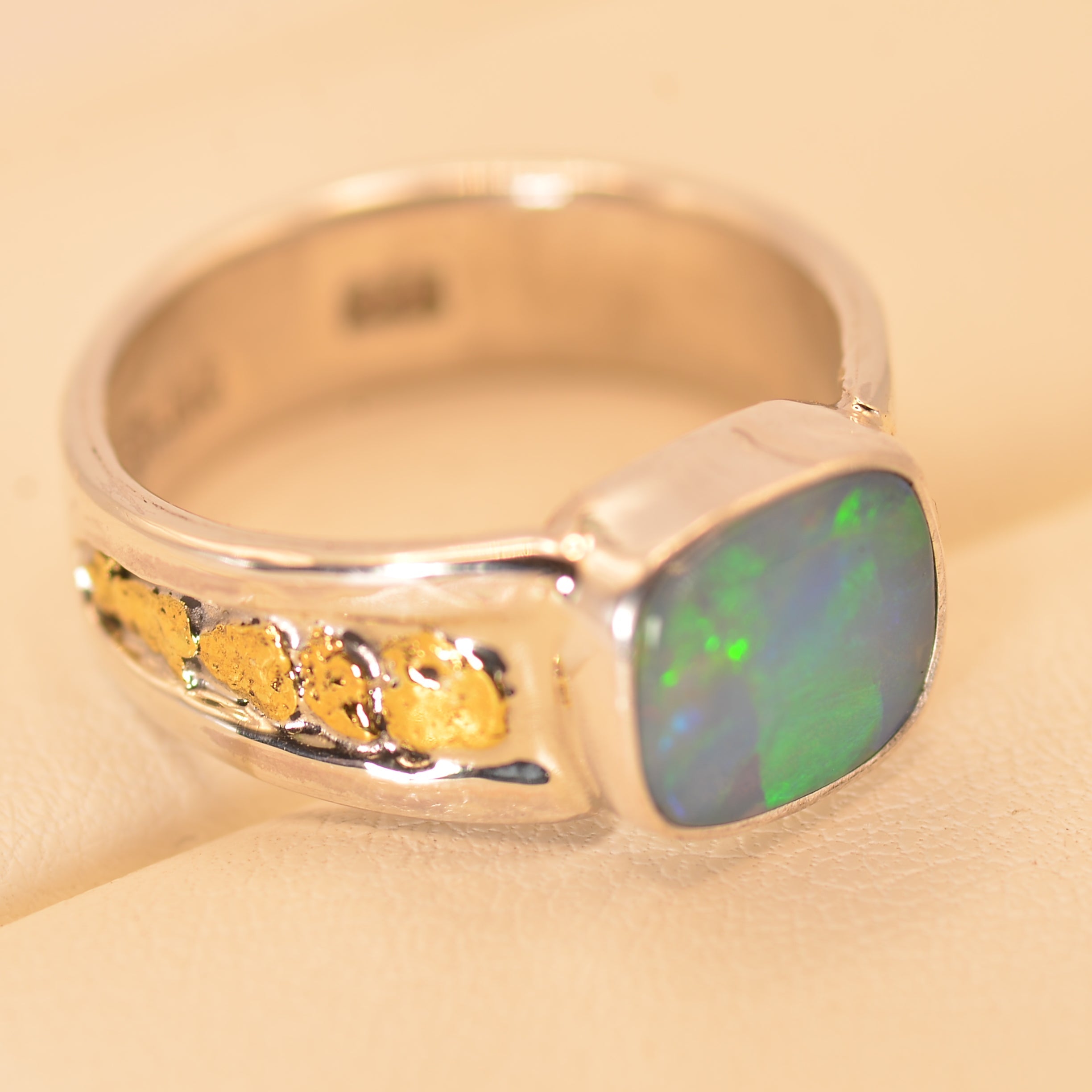 A silver ring featuring a square cabochon opal with green and blue play-of-color, accented by small yellow gemstones on the band, set against a light beige background.