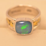A silver band ring featuring a square cabochon opal gemstone with a blue-gray body and a vibrant green flash, accented with small yellow inlays on the sides, set against a warm beige background.