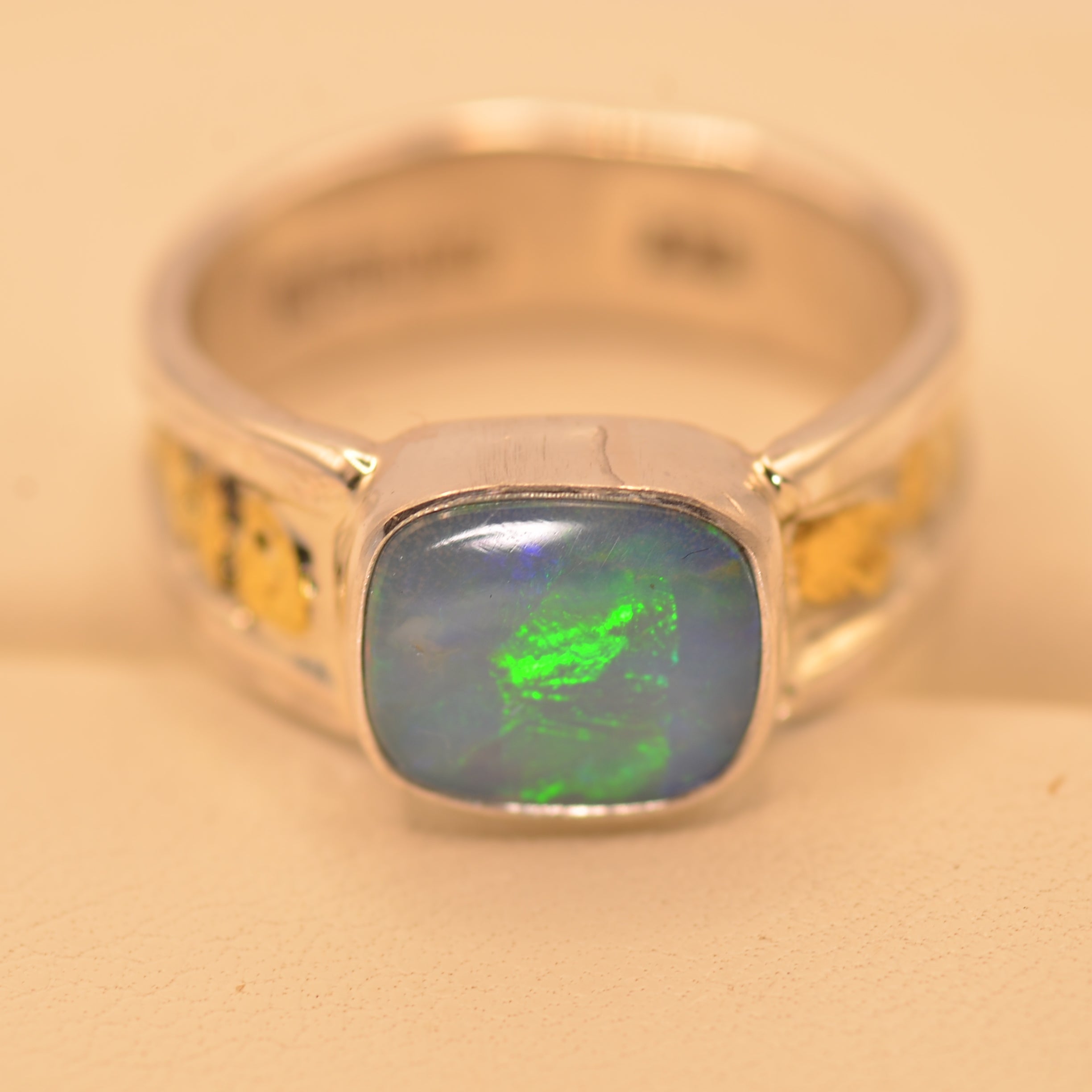 A silver band ring featuring a square cabochon opal gemstone with a blue-gray body and a vibrant green flash, accented with small yellow inlays on the sides, set against a warm beige background.