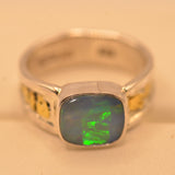 A silver ring featuring a square cabochon-cut opal stone with vibrant green and blue play-of-color, accented with small yellow gemstones on the shoulders, set against a plain beige background.