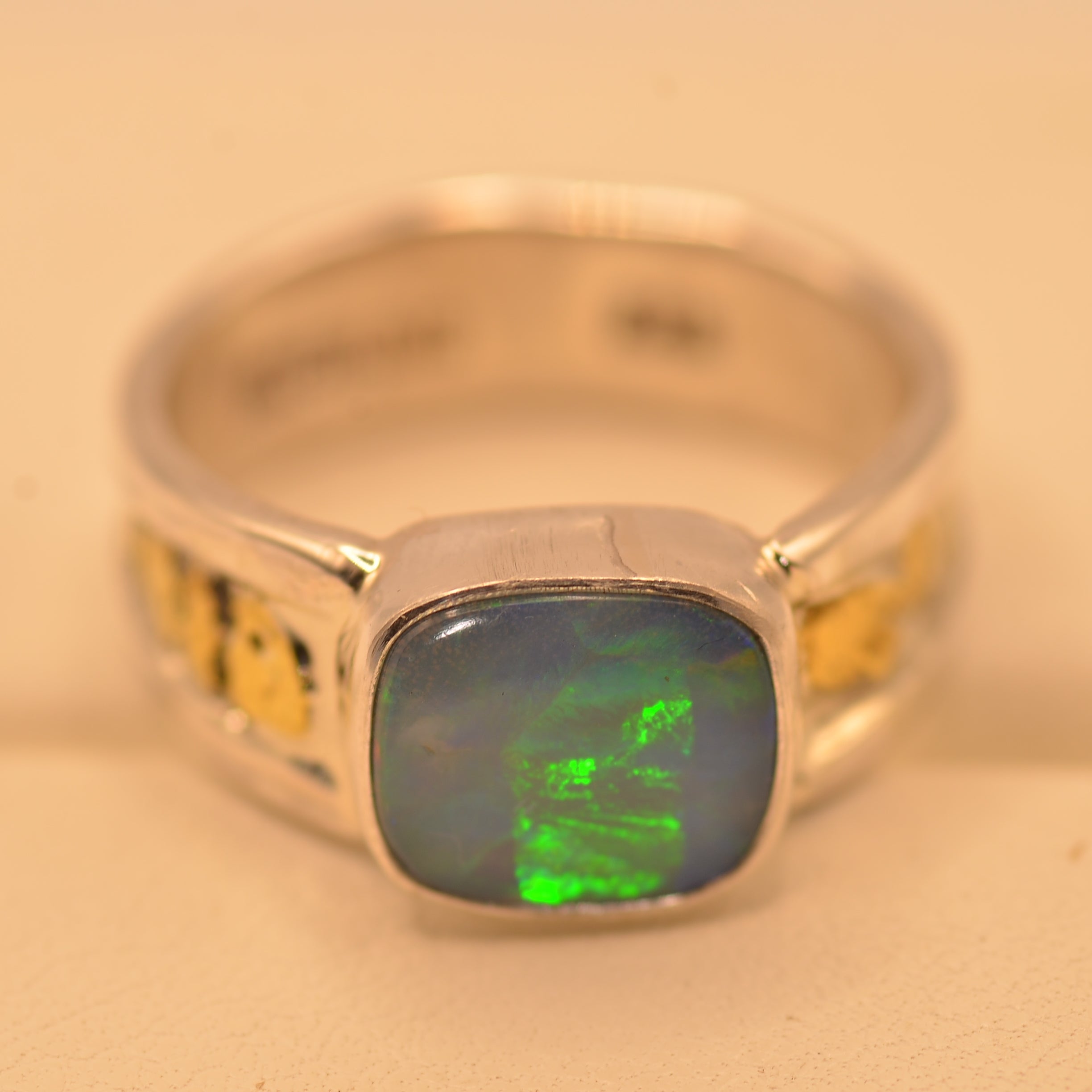 A silver ring featuring a square cabochon-cut opal stone with vibrant green and blue play-of-color, accented with small yellow gemstones on the shoulders, set against a plain beige background.