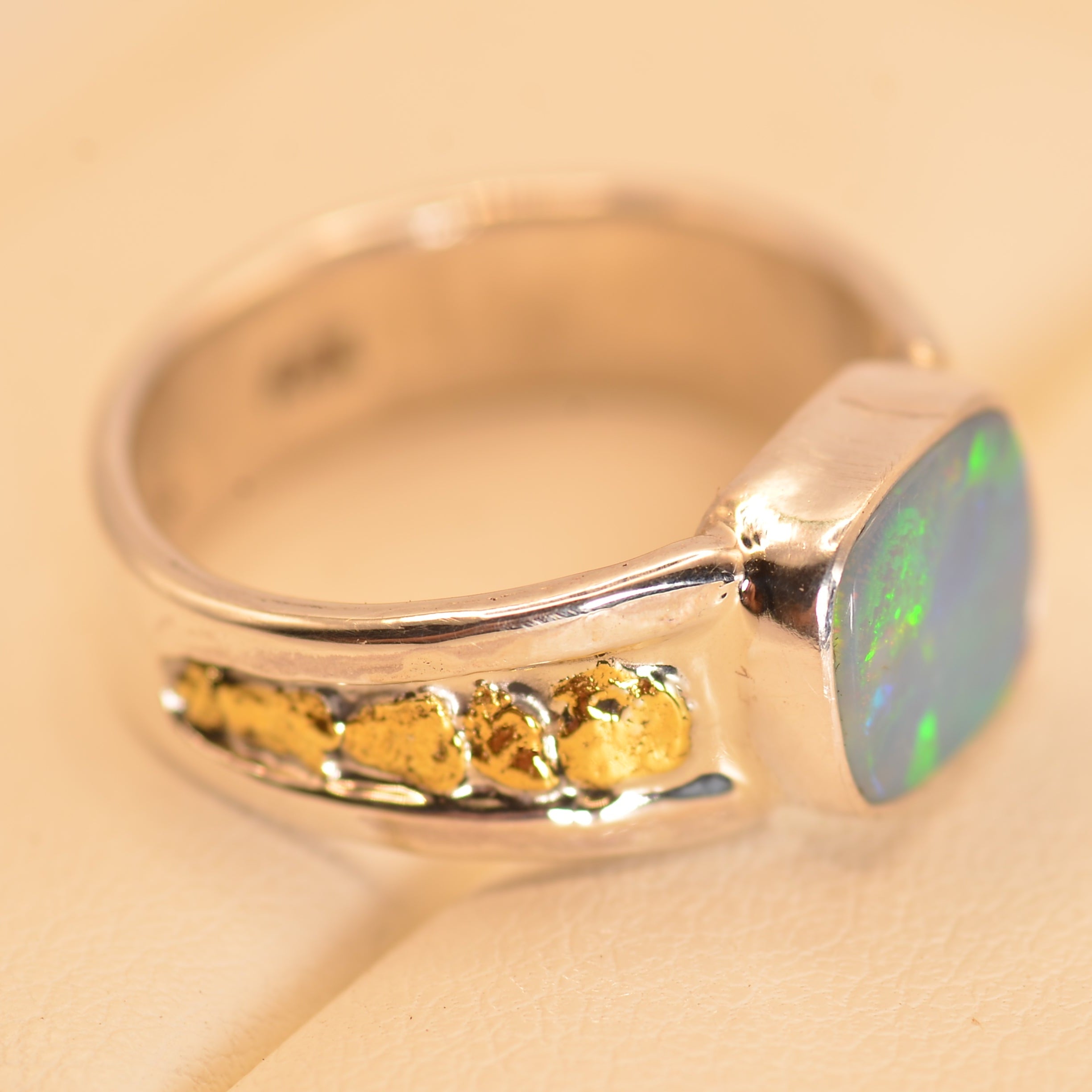 A silver ring with a square iridescent blue-green opal stone and a row of small gold inlays on the band, shown on a beige surface.