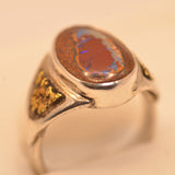 A silver ring featuring a large oval cabochon opal stone with a reddish-brown center and vivid blue iridescent flashes, accented with small gold inlays on the shoulders, set against a plain light beige background.