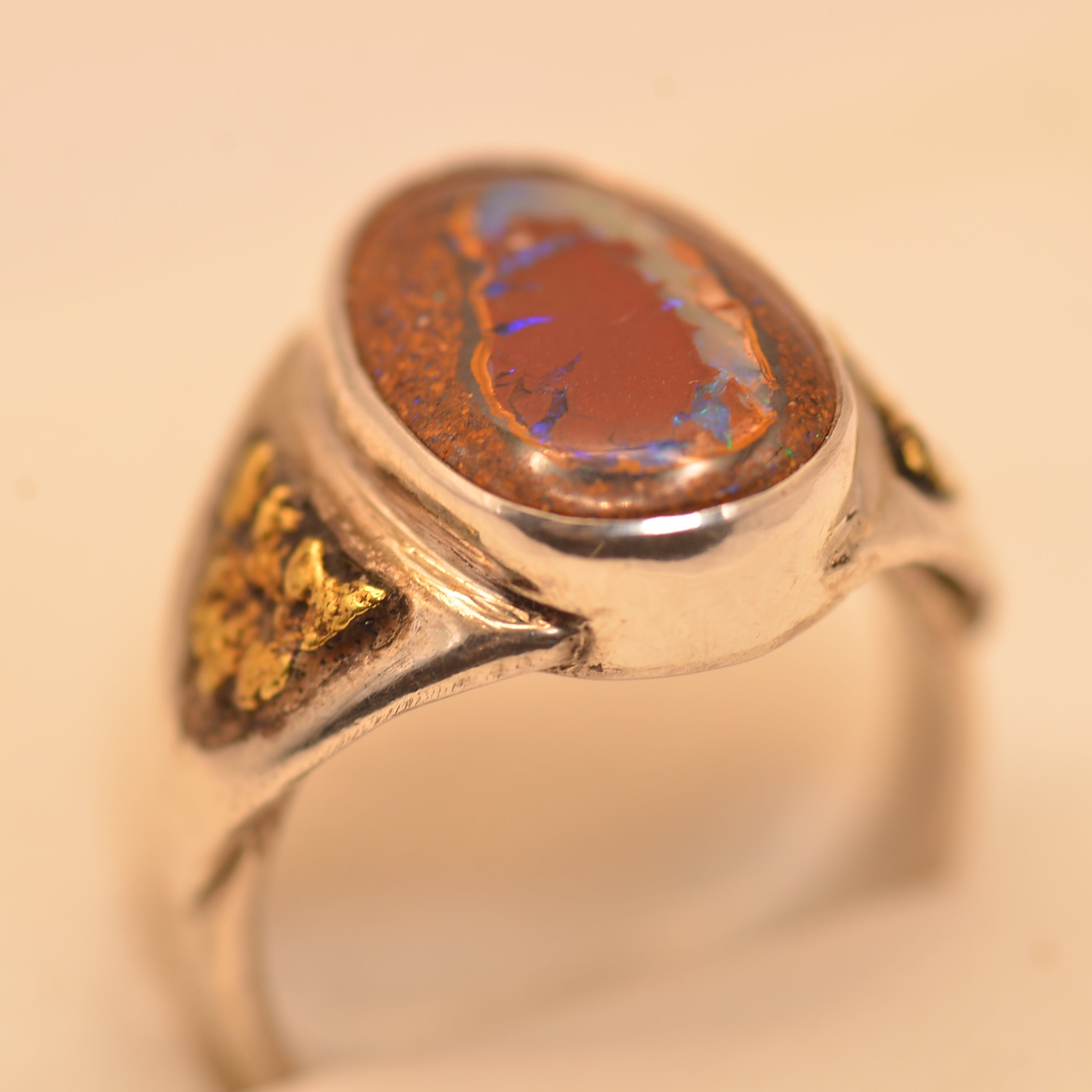 A silver ring featuring a large oval cabochon opal stone with a reddish-brown center and vivid blue iridescent flashes, accented with small gold inlays on the shoulders, set against a plain light beige background.