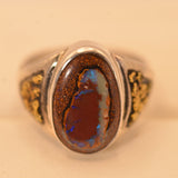 A silver-toned ring featuring a large oval brown opal stone with vibrant blue and orange flashes, set against a warm beige background.