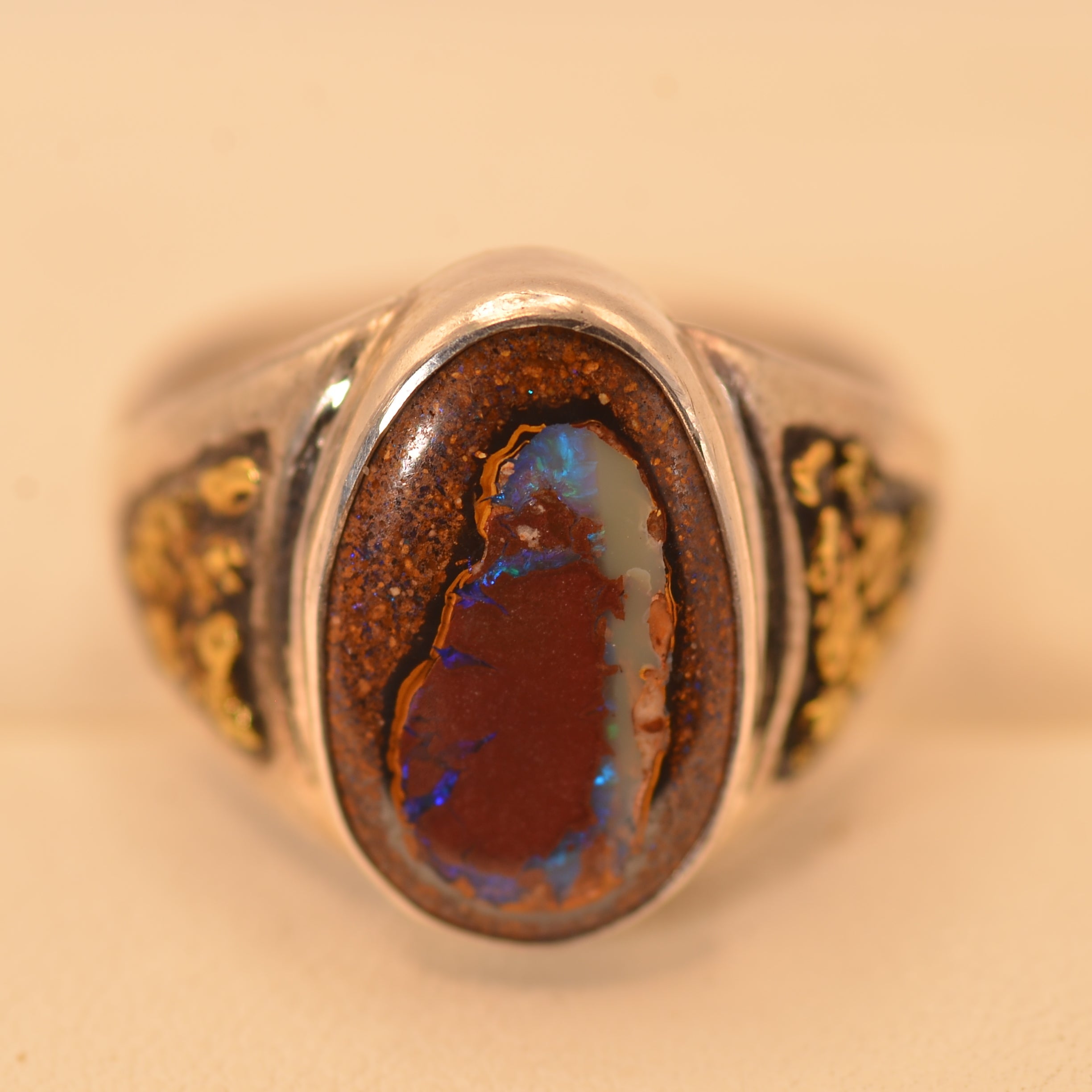 A silver-toned ring featuring a large oval brown opal stone with vibrant blue and orange flashes, set against a warm beige background.
