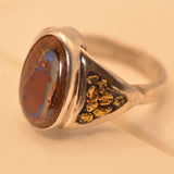 A sterling silver signet ring featuring a round brown and blue opal stone on one side and textured gold inlay on the other, resting on a beige surface.