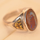 A silver ring featuring a large oval reddish-brown opal stone with iridescent blue flashes and a triangular inset filled with gold flakes, shown on a beige background.