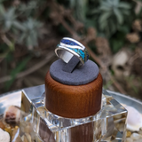 Silver ring with blue and green inlay on a wooden and fabric stand, surrounded by natural elements.