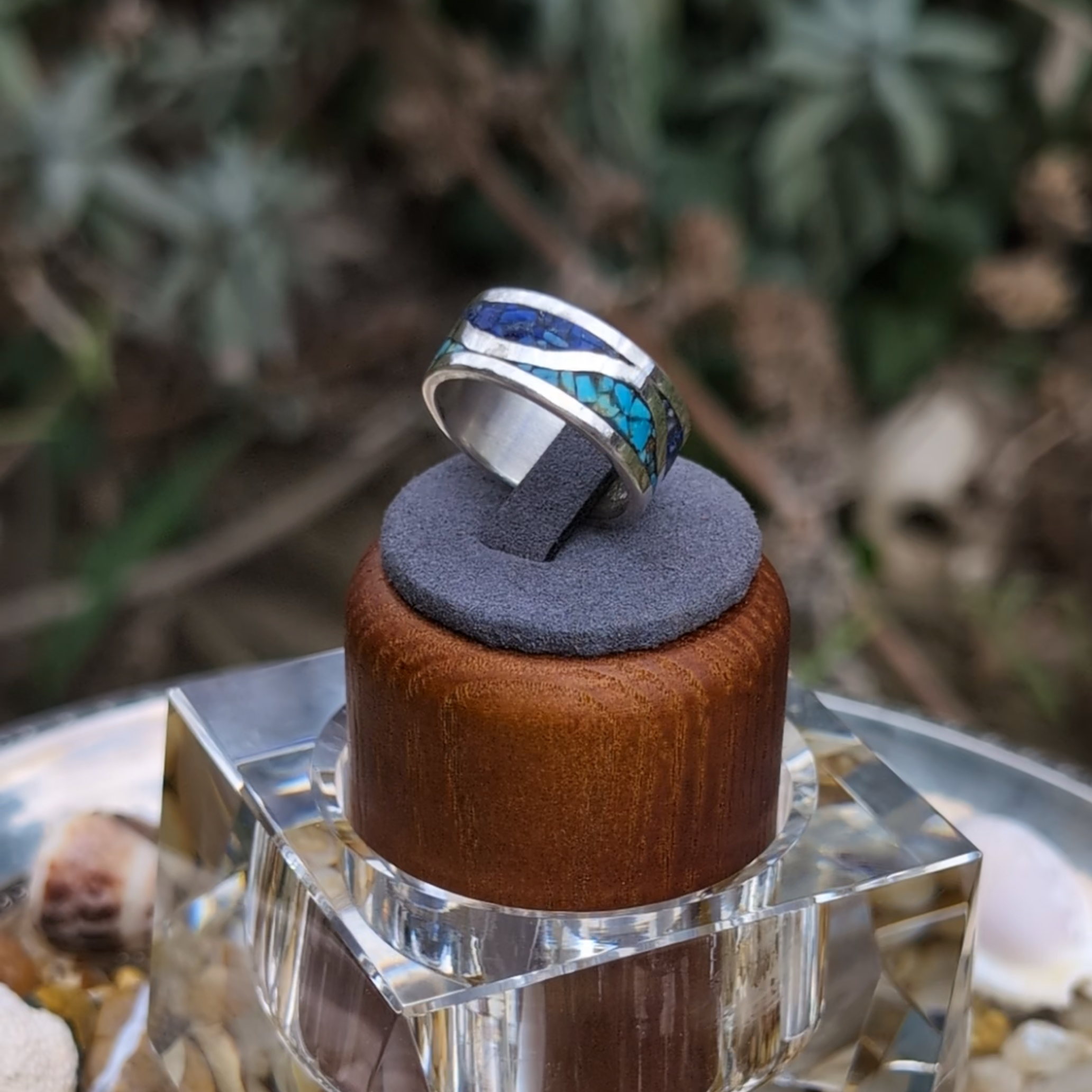 Silver ring with blue and green inlay on a wooden and fabric stand, surrounded by natural elements.
